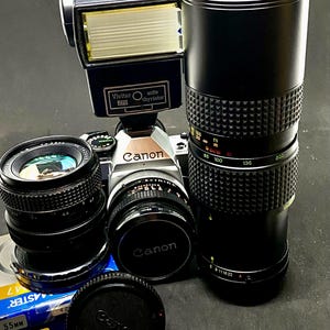 May include: A collection of vintage Canon camera equipment, including a silver and black Canon camera body, multiple lenses, a Vivitar 273 flash, and a lens cap. The lenses vary in size and focal length, with one labeled 55mm.