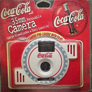 May include: A red and white 35mm reusable camera with a Coca-Cola logo on the front. The camera is packaged in a clear plastic clamshell with a red background and a Coca-Cola logo.