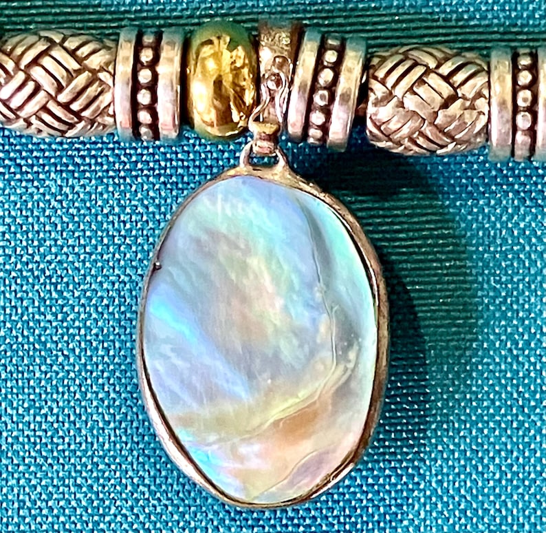 May include: A necklace featuring an oval pendant with an iridescent, pearlescent surface set in a silver-toned frame. The chain consists of alternating silver-toned beads with woven textures, smooth cylindrical spacers, and a single gold-toned bead.