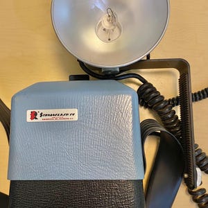 May include: Vintage StroboFlash IV camera flash unit with a silver reflector and a light blue and black body. The unit has a black cord and a label that reads "StroboFlash IV, Graflex Inc., Rochester, N.Y."