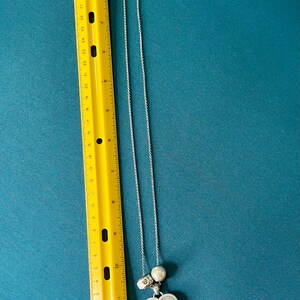 May include: A silver-toned necklace with a heart-shaped pendant featuring engraved text, a round white bead, and a small metallic charm, positioned next to a yellow ruler showing a length of approximately 12 inches for the chain and pendant.