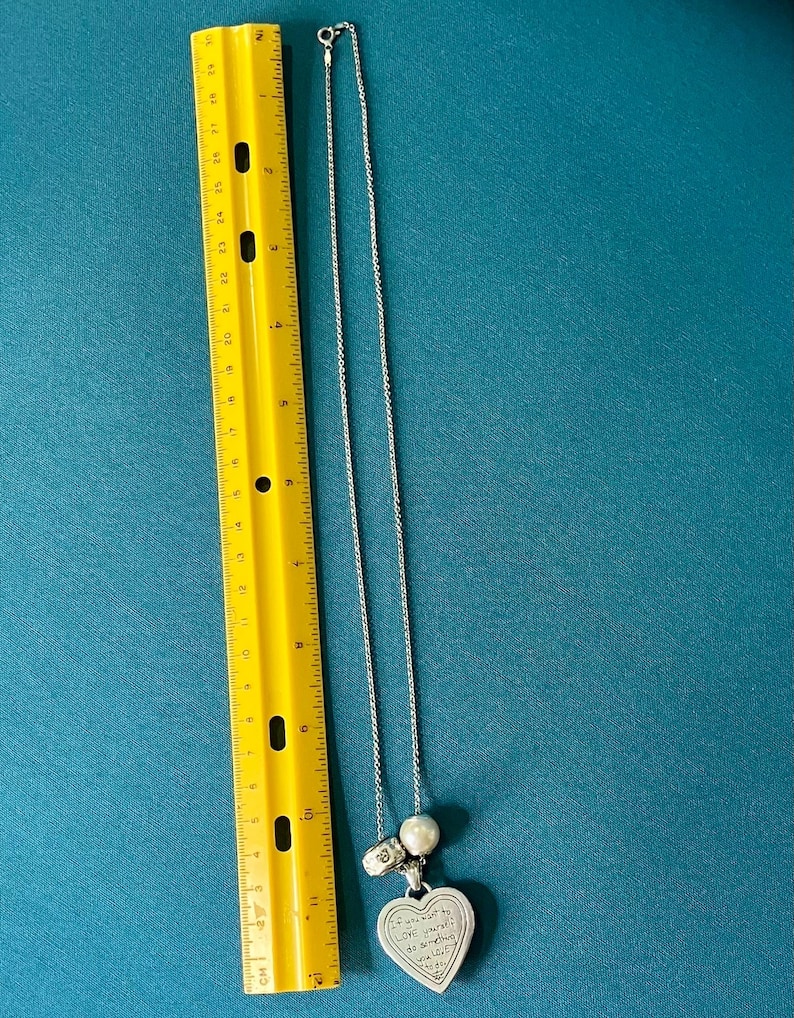 May include: A silver-toned necklace with a heart-shaped pendant featuring engraved text, a round white bead, and a small metallic charm, positioned next to a yellow ruler showing a length of approximately 12 inches for the chain and pendant.