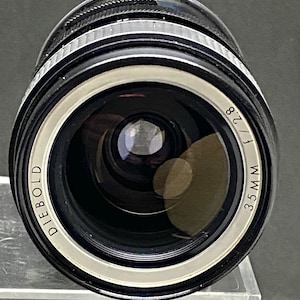 May include: A black and silver Diebold 35mm f/2.8 camera lens. The lens has a white ring with the Diebold brand name and the focal length and aperture details. The lens is cylindrical with a glass element.
