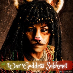 Pode incluir: Uma mulher vestida como a deusa egípcia da guerra Sekhmet. Ela tem maquiagem de gato, uma peruca preta com contas douradas e um colar de contas. O texto "War Goddess Sekhmet" está escrito em dourado sobre um fundo vermelho.