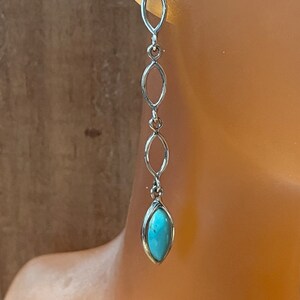May include: Silver-toned dangle earrings with a turquoise-colored stone at the bottom. The earrings feature three elongated oval links connected by small rings. The top link is a single oval shape. The earrings are displayed on a mannequin.