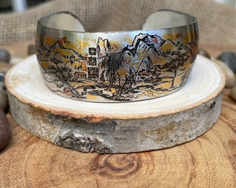 May include: Silver-toned cuff bracelet with a landscape design. The cuff features a detailed illustration of mountains, trees, and buildings in shades of black, gold, and orange. The bracelet is displayed on a wooden slice.