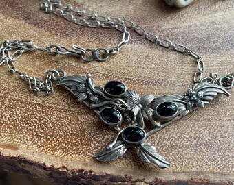 May include: A silver-toned necklace with a floral design, featuring five oval black gemstones. The necklace has a chain and a decorative pendant with leaves and flowers. The jewelry is displayed on a wooden surface.