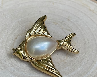 May include: A gold-toned fish brooch with a pearlescent, teardrop-shaped pearl at its center. The brooch features detailed fins and tail, and is set against a light-colored wooden surface. This jewelry piece is a unique accessory.