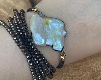 May include: A pearl bracelet with a large, irregularly shaped white pearl pendant. The pendant is attached to a dark brown cord bracelet with gold-colored accents. A separate wrap bracelet with dark, faceted beads is also visible.