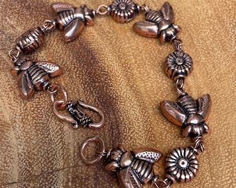 May include: Copper-toned bracelet featuring bee and flower charms. The bracelet has a lobster clasp closure and is displayed on a wooden surface. The bee charms have detailed wings and body segments, while the flower charms have a detailed petal design.