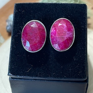 May include: A pair of oval-shaped ruby earrings with a faceted cut, set in a silver bezel. The earrings are displayed on a black velvet jewelry box, showcasing their vibrant red color and sparkle. A perfect gift for any occasion.