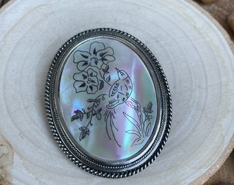 May include: Oval silver-toned brooch with a mother-of-pearl center. The center features a detailed engraving of a bird perched on a branch with flowers. The brooch has a twisted rope-like border and is set on a wooden surface.