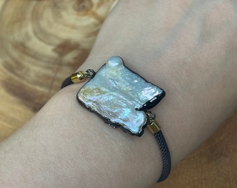 May include: A bracelet featuring a large, irregularly shaped, iridescent pearl pendant with a dark border. The pearl is attached to a dark gray cord with gold-colored metal accents.