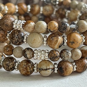May include: Close-up of several stacked bracelets. Each bracelet features round, speckled, earth-toned beads, interspersed with small silver-colored spacers. The beads vary in color, from light beige to dark brown. The bracelets are stacked, creating a layered effect.