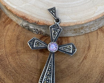 May include: A silver-toned cross pendant with a central, round, light purple gemstone. The cross features a detailed design with small, dark accents and a triangular top loop for a chain. The pendant is displayed on a wooden surface.