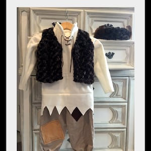 May include: A brown faux fur waistcoat with a white shirt and brown trousers. The waistcoat has a lace-up front and the trousers have a patch on the knee. The outfit is hanging on a hanger in front of a white dresser.