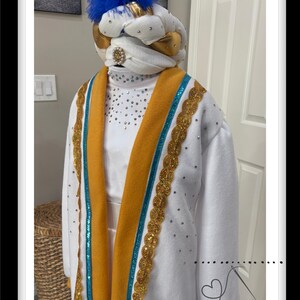 Peut inclure: Robe blanche avec une bordure dorée et turquoise, ornée d'un turban blanc surmonté d'une plume bleue. La robe est dotée d'une ceinture dorée et est décorée de paillettes.