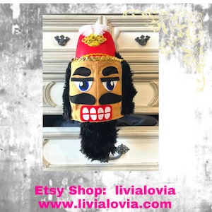 Puede incluir: Una cabeza de cascanueces caprichosa con un sombrero rojo, adornos dorados y una cara color canela. La cara tiene ojos azules, un bigote negro y una boca roja y blanca. Piel negra rodea la cara. El nombre y el sitio web de la tienda Etsy son visibles.