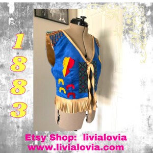 May include: A blue sleeveless vest with a geometric pattern and fringe trim. The vest has a tie closure and is decorated with colorful patches. The vest is on a mannequin.