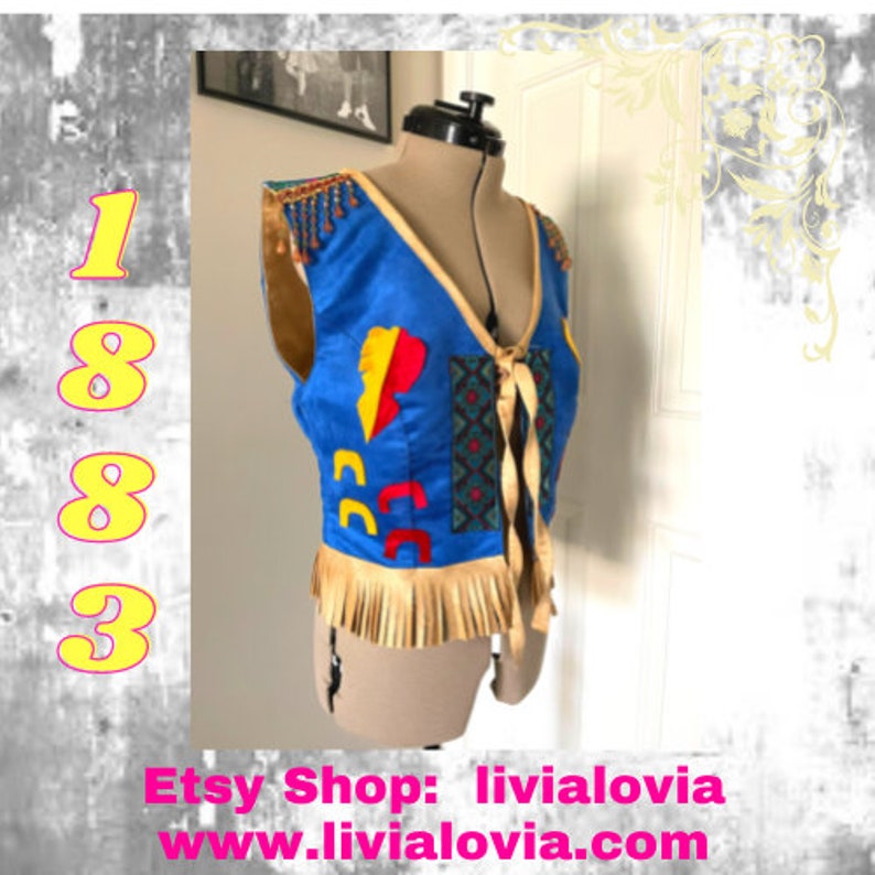May include: A blue sleeveless vest with a geometric pattern and fringe trim. The vest has a tie closure and is decorated with colorful patches. The vest is on a mannequin.