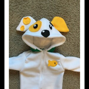 May include: A white dog costume with a hood featuring yellow and black accents, a green collar, and a yellow fish applique. The costume has a zipper closure and is designed for a child.