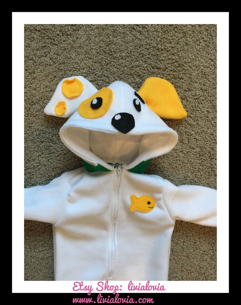 May include: A white dog costume with a hood featuring yellow and black accents, a green collar, and a yellow fish applique. The costume has a zipper closure and is designed for a child.