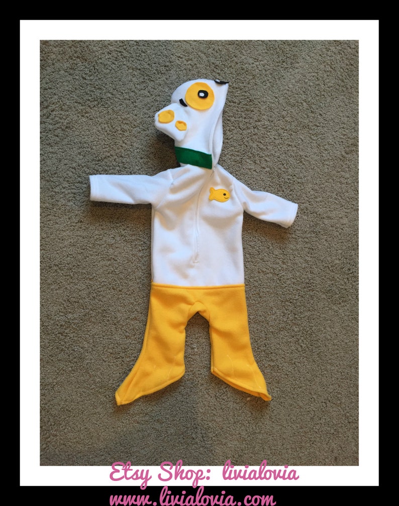 May include: A white and yellow baby outfit resembling a duck. The hooded jumpsuit features a duck head with yellow eyes and beak, a green collar, and a small yellow fish applique. The bottom half is yellow.