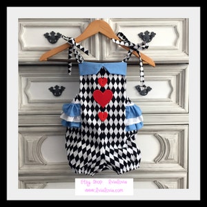 May include: A black and white diamond patterned baby onesie with a blue collar and red felt hearts. The onesie has white ruffles on the shoulders and is hanging on a wooden hanger.