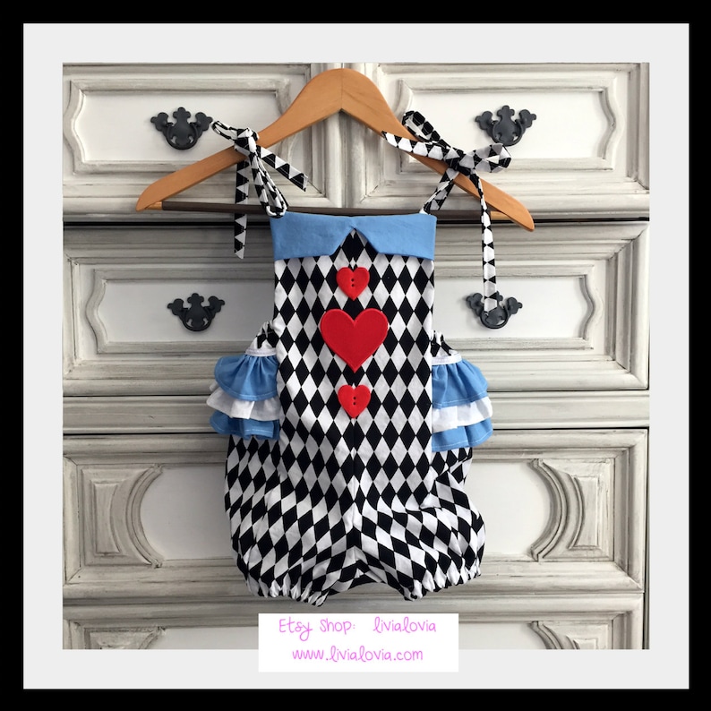 May include: A black and white diamond patterned baby onesie with a blue collar and red felt hearts. The onesie has white ruffles on the shoulders and is hanging on a wooden hanger.