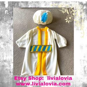 Peut inclure: Un ensemble de costume crème comprenant une robe avec une bordure jaune et une ceinture décorative avec des accents bleus et dorés. Un chapeau assorti avec une plume bleue complète la tenue. Le costume est pour enfants.