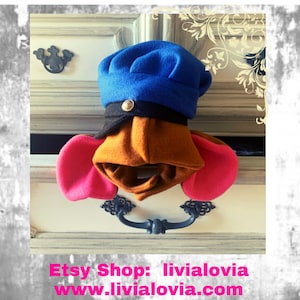 May include: A colorful, handmade hat and hood set in blue, black, orange, and pink. The hat has a blue top and a black brim with a gold button. The hood is orange with pink ear-like extensions. The set is displayed on a white drawer.