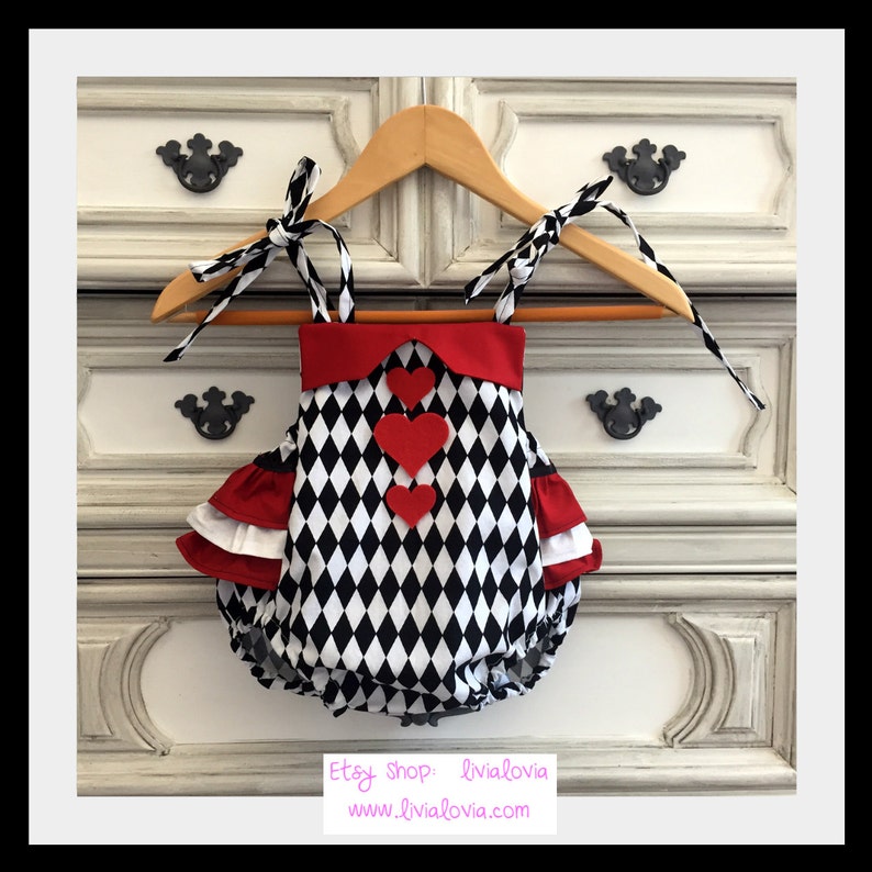 May include: A black and white diamond patterned baby onesie with red trim and three red felt hearts. The onesie has ruffled sleeves and ties at the shoulders.