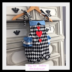 May include: A black and white diamond patterned baby onesie with a blue ruffled collar and red felt hearts. The onesie has straps that tie at the shoulders.