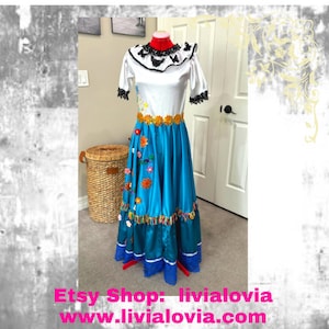 Puede incluir: Un vestido tradicional vibrante con una parte superior blanca y una falda azul brillante. La parte superior tiene ribetes de encaje negro y un cuello con volantes. La falda está adornada con aplicaciones florales coloridas y adornos. El nombre y el sitio web de la tienda Etsy son visibles en la parte inferior.