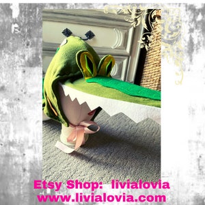 May include: A green felt alligator hat with yellow and green details, and a white, jagged mouth. The hat is designed to be worn on the head. The Etsy shop name and website are visible at the bottom of the image.