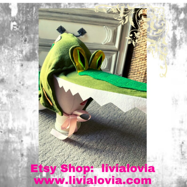 May include: A green felt alligator hat with yellow and green details, and a white, jagged mouth. The hat is designed to be worn on the head. The Etsy shop name and website are visible at the bottom of the image.