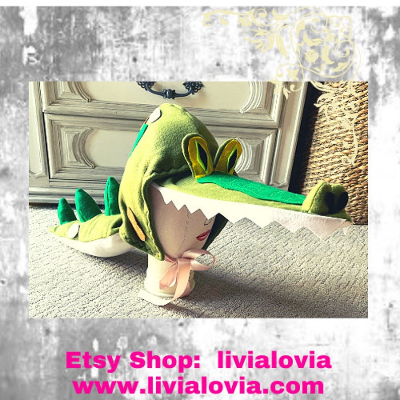 May include: A green alligator costume hat with white teeth, green eyes, and yellow and green ears. The hat is made of soft fabric and has a long tail. The Etsy shop name and website are displayed in pink text at the bottom.