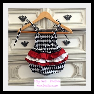 May include: A black and white diamond patterned baby onesie with a red and white ruffled skirt. The onesie has thin straps and is hanging on a wooden hanger.