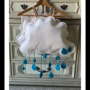 May include: A white felt cloud mobile with blue raindrop details suspended from a wooden hanger with rainbow straps. The mobile is designed for hanging. The Etsy shop name and website are visible at the bottom of the image.