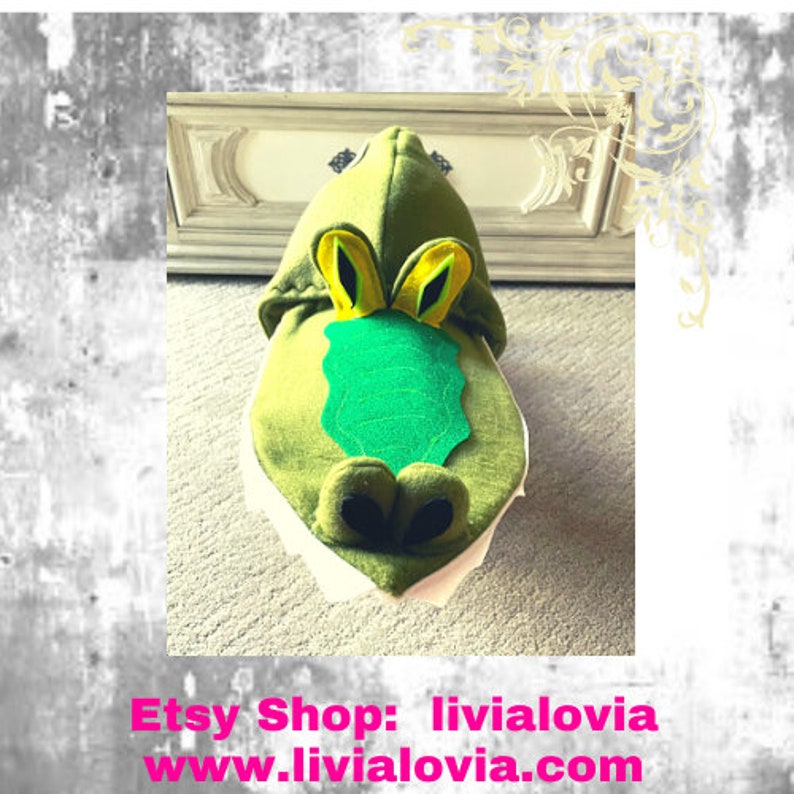 May include: A green alligator-themed hooded costume with yellow and black eye details, a green felt snout, and white fabric around the mouth. The Etsy shop name and website are displayed in pink text at the bottom.