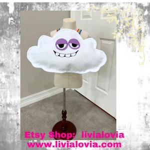 Puede incluir: Un disfraz blanco en forma de nube con una cara sonriente morada y tirantes arcoíris. El disfraz se muestra en un maniquí.