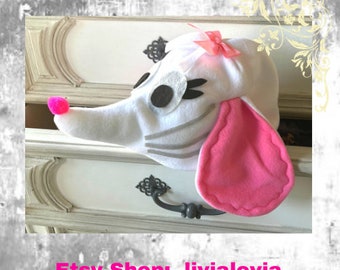 May include: A white felt dog hat with a pink nose, pink ear, and a pink bow. The hat has gray stripes and black and white eyes. The Etsy shop name and website are printed in pink at the bottom of the image.