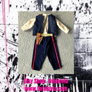 May include: A doll-sized outfit featuring a cream-colored shirt, a navy blue vest with a geometric pattern, and navy blue pants with red stripes. A brown holster and a gold-colored belt complete the ensemble. The outfit is displayed on a textured surface.