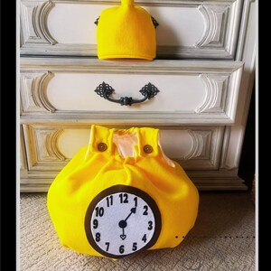 Puede incluir: Una bolsa de fieltro amarillo brillante con un diseño de esfera de reloj, con una esfera de reloj blanca con números y manecillas negras. Arriba, se muestra un sombrero de fieltro amarillo a juego. La bolsa tiene dos cierres de botón marrones. El fondo incluye una cómoda blanca y gris.