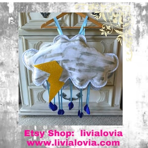 Puede incluir: Móvil con forma de nube con un rayo dorado brillante y detalles de gotas de lluvia azules. La nube es blanca con detalles de pinceladas grises. Cuelga de una percha de madera con correas azules. Texto: "Etsy Shop: livialovia www.livialovia.com".