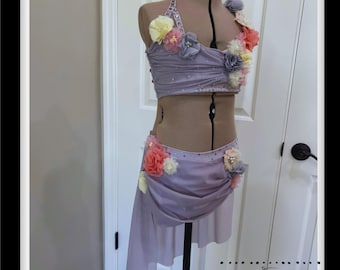 May include: A lavender dance costume on a mannequin. The two-piece outfit features a halter top and a skirt, both adorned with floral embellishments. The top and skirt are embellished with sequins. The costume is displayed in front of a white door.