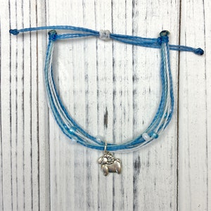 Op de afbeelding: Gevlochten armband in blauw- en wittinten met een zilveren ram-bedel. De armband heeft meerdere strengen blauw en wit koord, met kleine witte kralen. De bedel is een gedetailleerde ram.