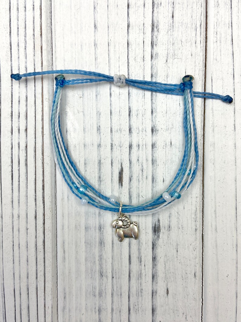 Op de afbeelding: Gevlochten armband in blauw- en wittinten met een zilveren ram-bedel. De armband heeft meerdere strengen blauw en wit koord, met kleine witte kralen. De bedel is een gedetailleerde ram.