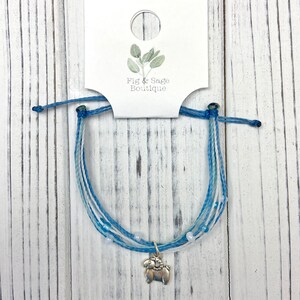 Op de afbeelding: Een blauw-witte vriendschapsarmband met een zilveren bedel van een ram. De armband is gemaakt van meerdere strengen blauw en wit koord, met kleine witte kralen. Het label toont de tekst "Fig & Sage Boutique".
