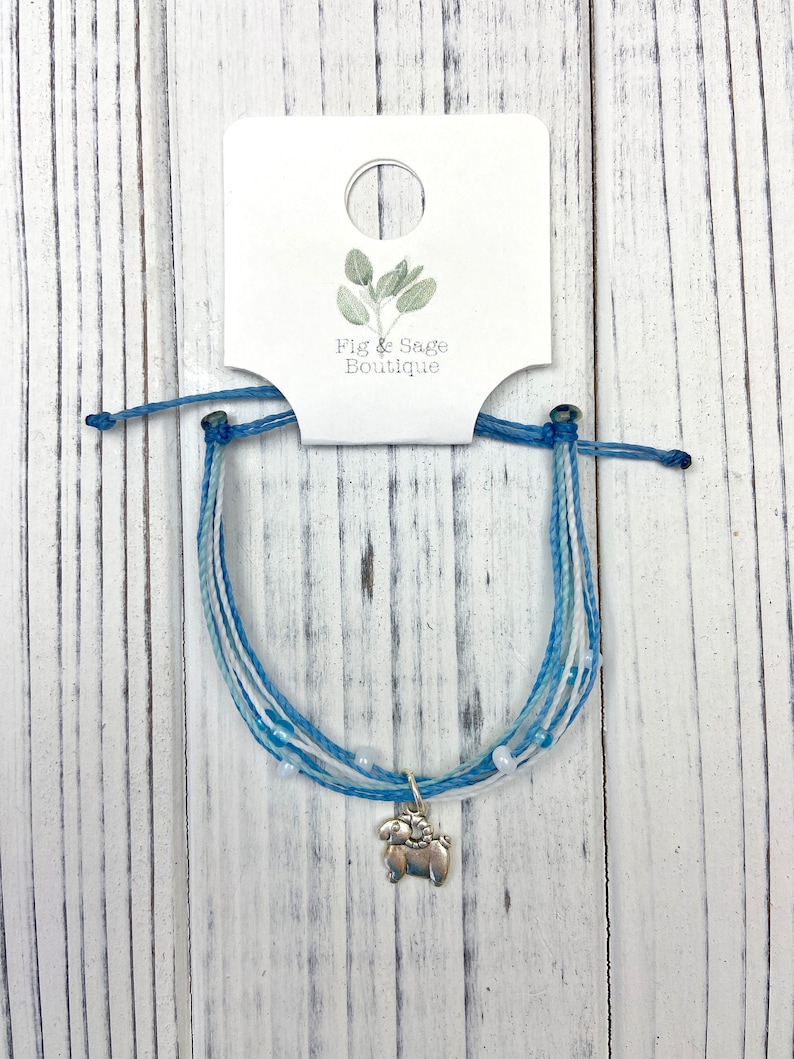 Op de afbeelding: Een blauw-witte vriendschapsarmband met een zilveren bedel van een ram. De armband is gemaakt van meerdere strengen blauw en wit koord, met kleine witte kralen. Het label toont de tekst "Fig & Sage Boutique".